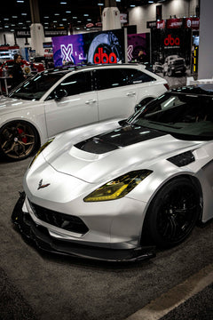 Silver Chevrolet Corvette race car displayed at an indoor automotive exhibition, featuring a wide-body aerodynamic kit, large rear wing, carbon fiber front splitter, and vented hood. The aggressive motorsport styling is showcased under exhibition lighting with REVOZPORT branding in the background.