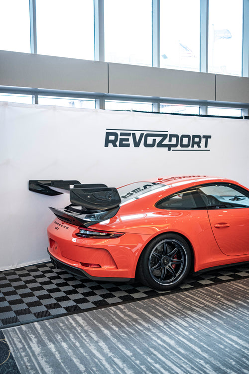 An orange Porsche sports car displayed indoors at an automotive exhibition booth, featuring an aggressive front splitter, black performance wheels, and a sleek aerodynamic body kit. The car is parked in front of a REVOZPORT branded display wall inside a modern convention center with large windows and exhibition setups in the background.