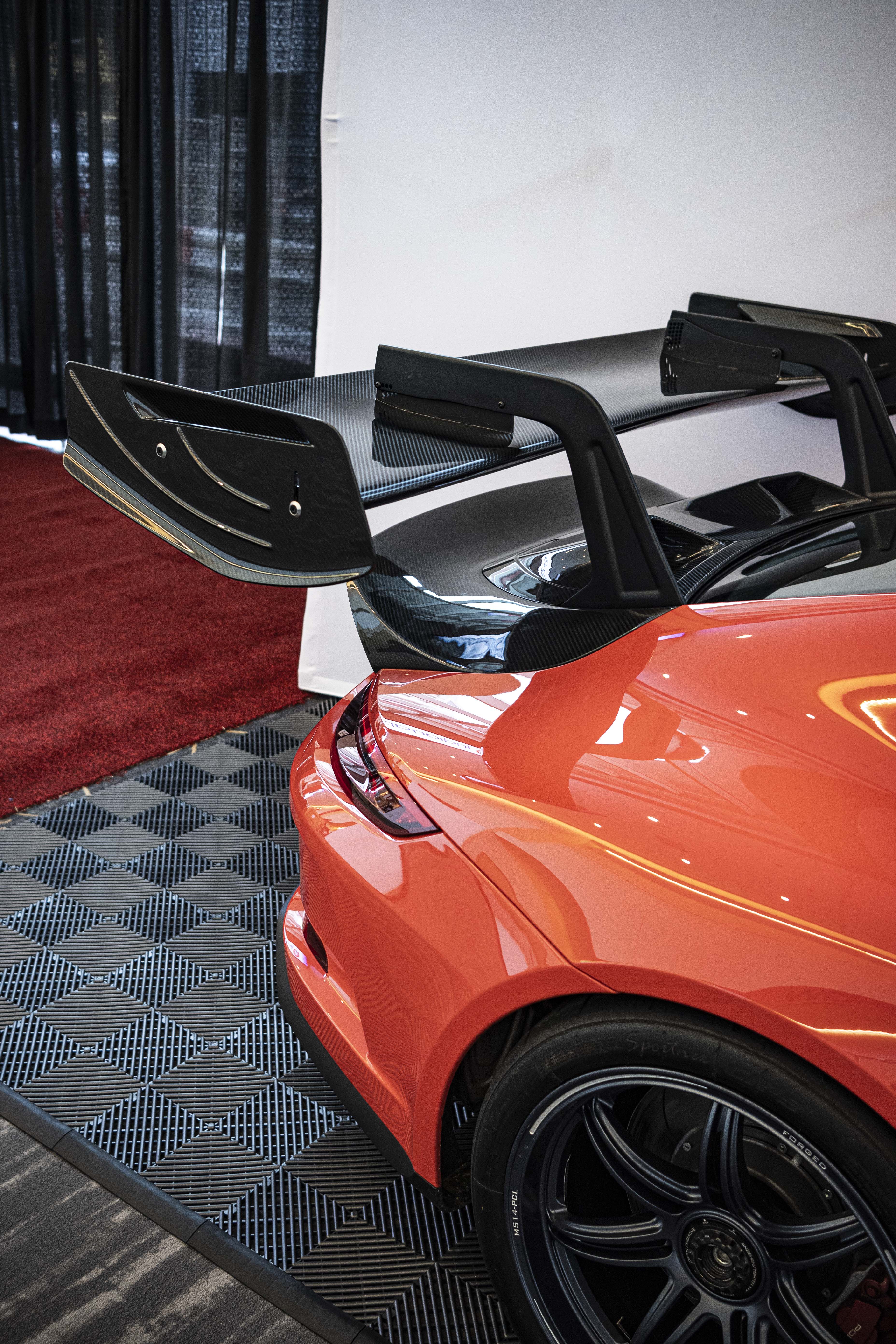 Close-up of a performance wheel setup on an orange Porsche sports car, featuring black multi-spoke forged rims, red brake calipers, and a large drilled brake rotor. The image highlights the aggressive fitment, low-profile tire, and carbon fiber side skirt details at an indoor automotive display.