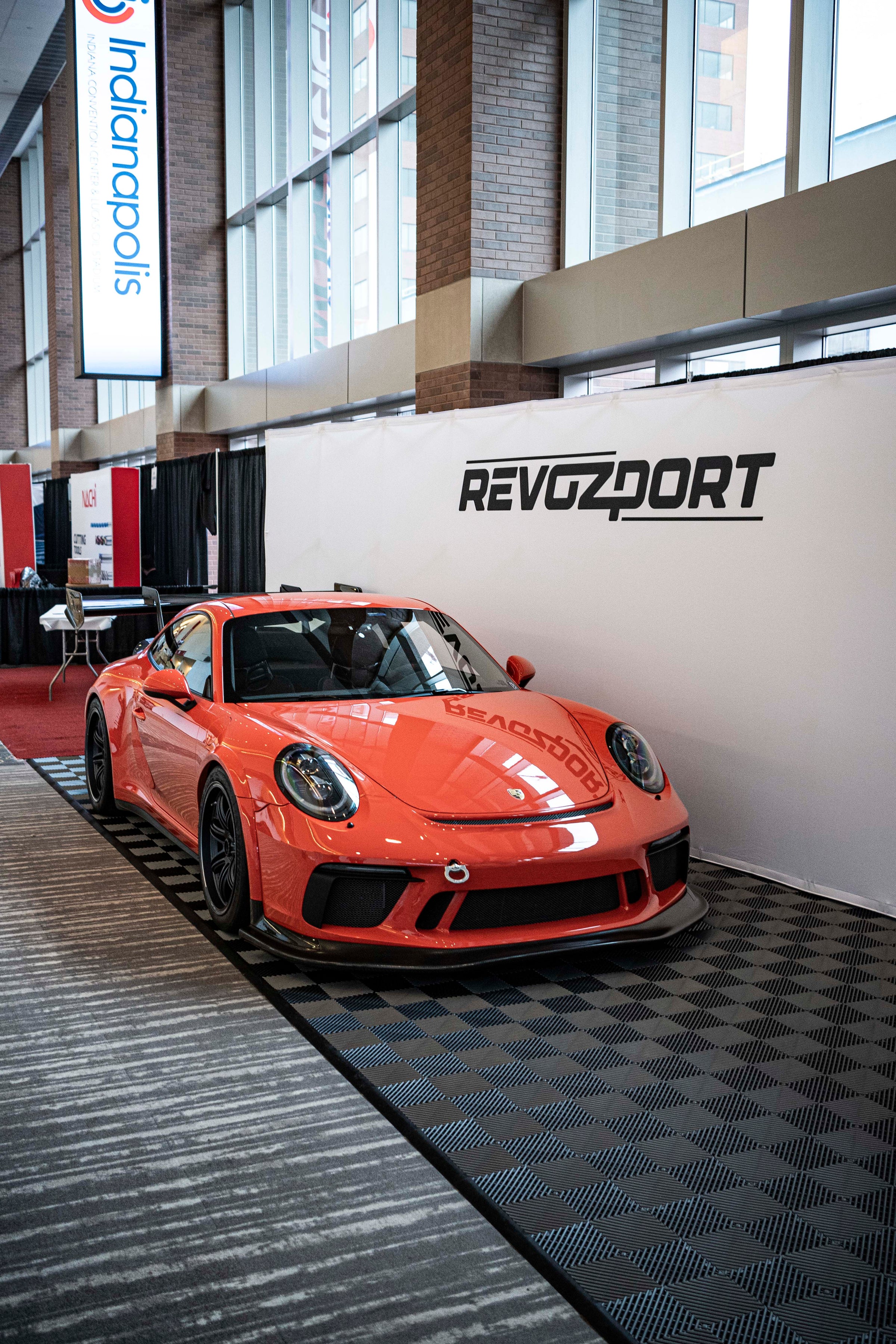 An orange Porsche sports car displayed indoors at an automotive exhibition booth, featuring an aggressive front splitter, black performance wheels, and a sleek aerodynamic body kit. The car is parked in front of a REVOZPORT branded display wall inside a modern convention center with large windows and exhibition setups in the background.