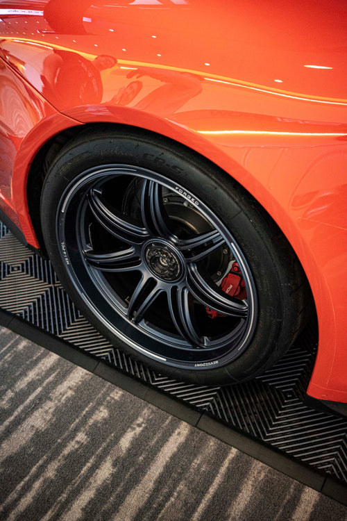 An orange Porsche sports car displayed indoors at an automotive exhibition booth, featuring an aggressive front splitter, black performance wheels, and a sleek aerodynamic body kit. The car is parked in front of a REVOZPORT branded display wall inside a modern convention center with large windows and exhibition setups in the background.