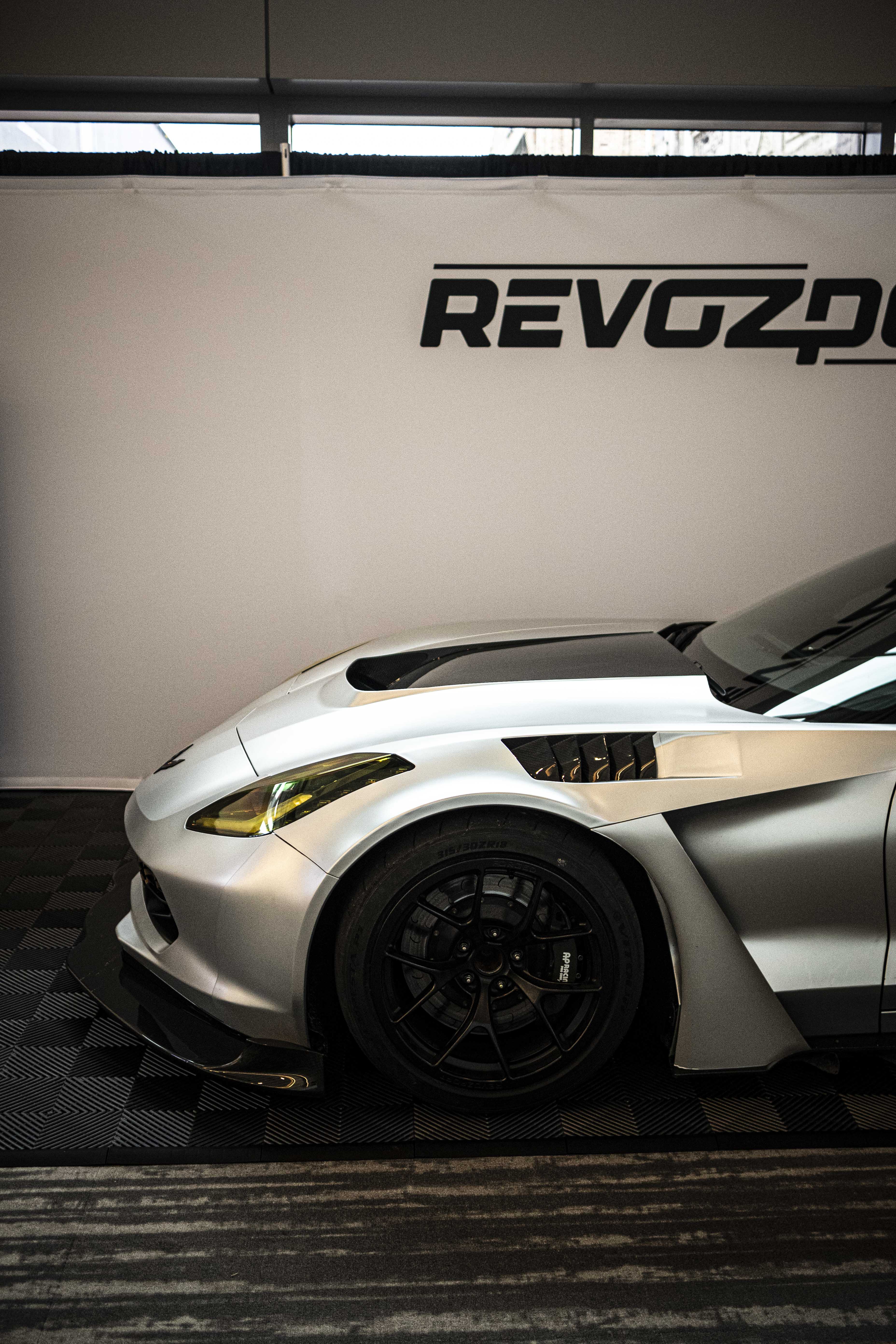 Silver Chevrolet Corvette race car displayed at an indoor automotive exhibition, featuring a wide-body aerodynamic kit, large rear wing, carbon fiber front splitter, and vented hood. The aggressive motorsport styling is showcased under exhibition lighting with REVOZPORT branding in the background.