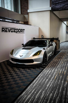 Silver Chevrolet Corvette race car displayed at an indoor automotive exhibition, featuring a wide-body aerodynamic kit, large rear wing, carbon fiber front splitter, and vented hood. The aggressive motorsport styling is showcased under exhibition lighting with REVOZPORT branding in the background.