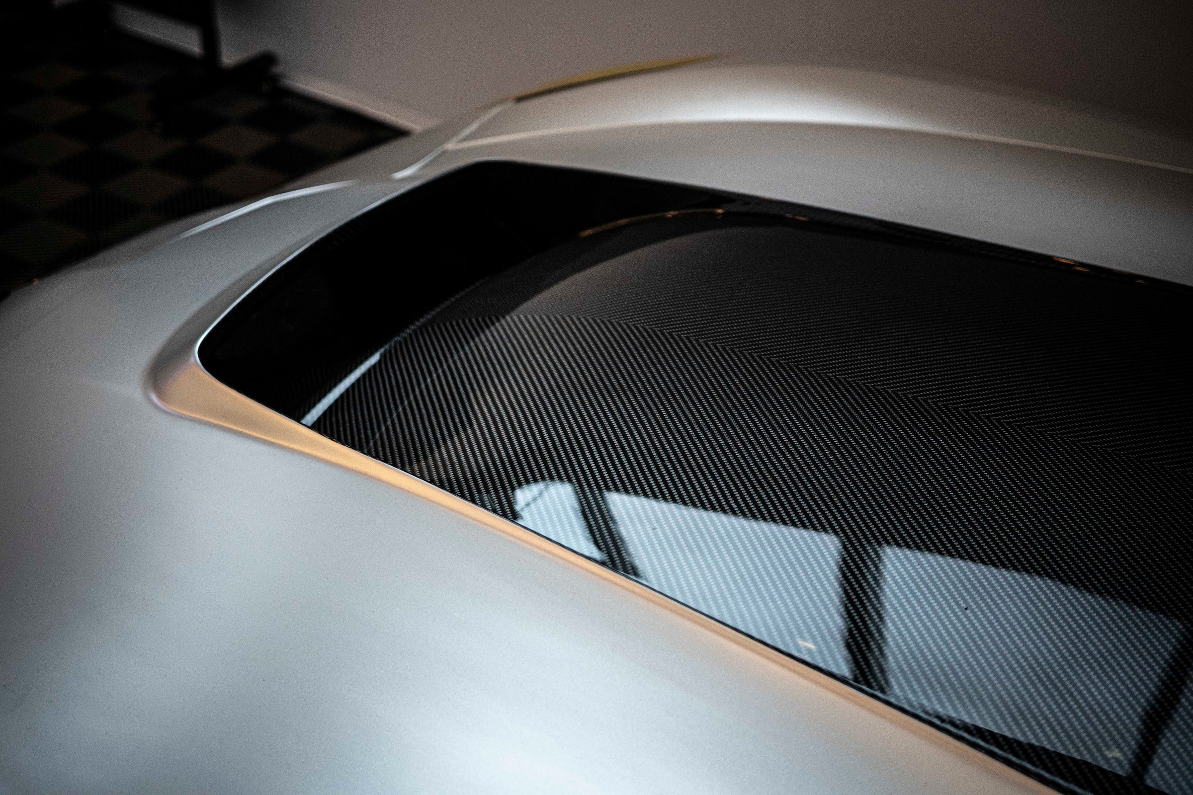 Silver Chevrolet Corvette race car displayed at an indoor automotive exhibition, featuring a wide-body aerodynamic kit, large rear wing, carbon fiber front splitter, and vented hood. The aggressive motorsport styling is showcased under exhibition lighting with REVOZPORT branding in the background.