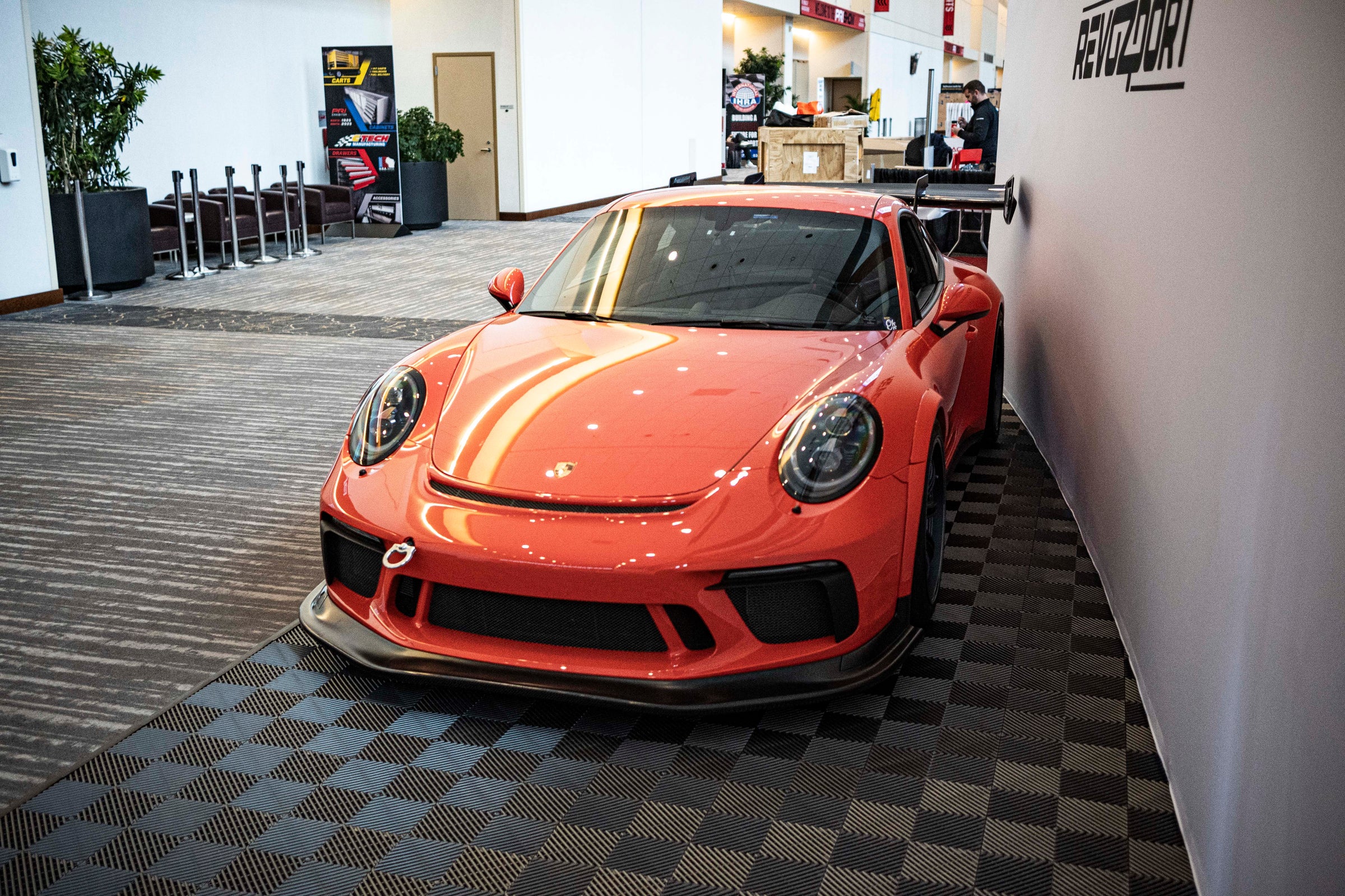 An orange Porsche sports car displayed indoors at an automotive exhibition booth, featuring an aggressive front splitter, black performance wheels, and a sleek aerodynamic body kit. The car is parked in front of a REVOZPORT branded display wall inside a modern convention center with large windows and exhibition setups in the background.