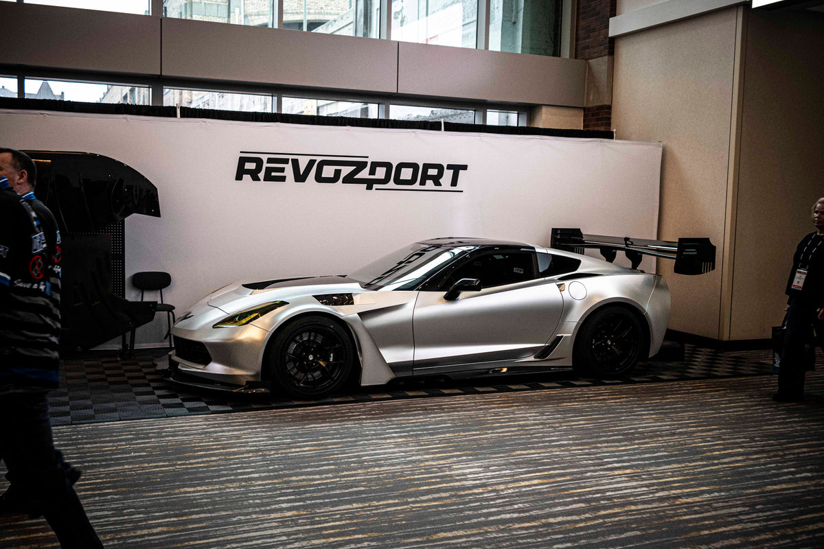 Silver Chevrolet Corvette race car displayed at an indoor automotive exhibition, featuring a wide-body aerodynamic kit, large rear wing, carbon fiber front splitter, and vented hood. The aggressive motorsport styling is showcased under exhibition lighting with REVOZPORT branding in the background.