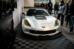 Silver Chevrolet Corvette race car displayed at an indoor automotive exhibition, featuring a wide-body aerodynamic kit, large rear wing, carbon fiber front splitter, and vented hood. The aggressive motorsport styling is showcased under exhibition lighting with REVOZPORT branding in the background.