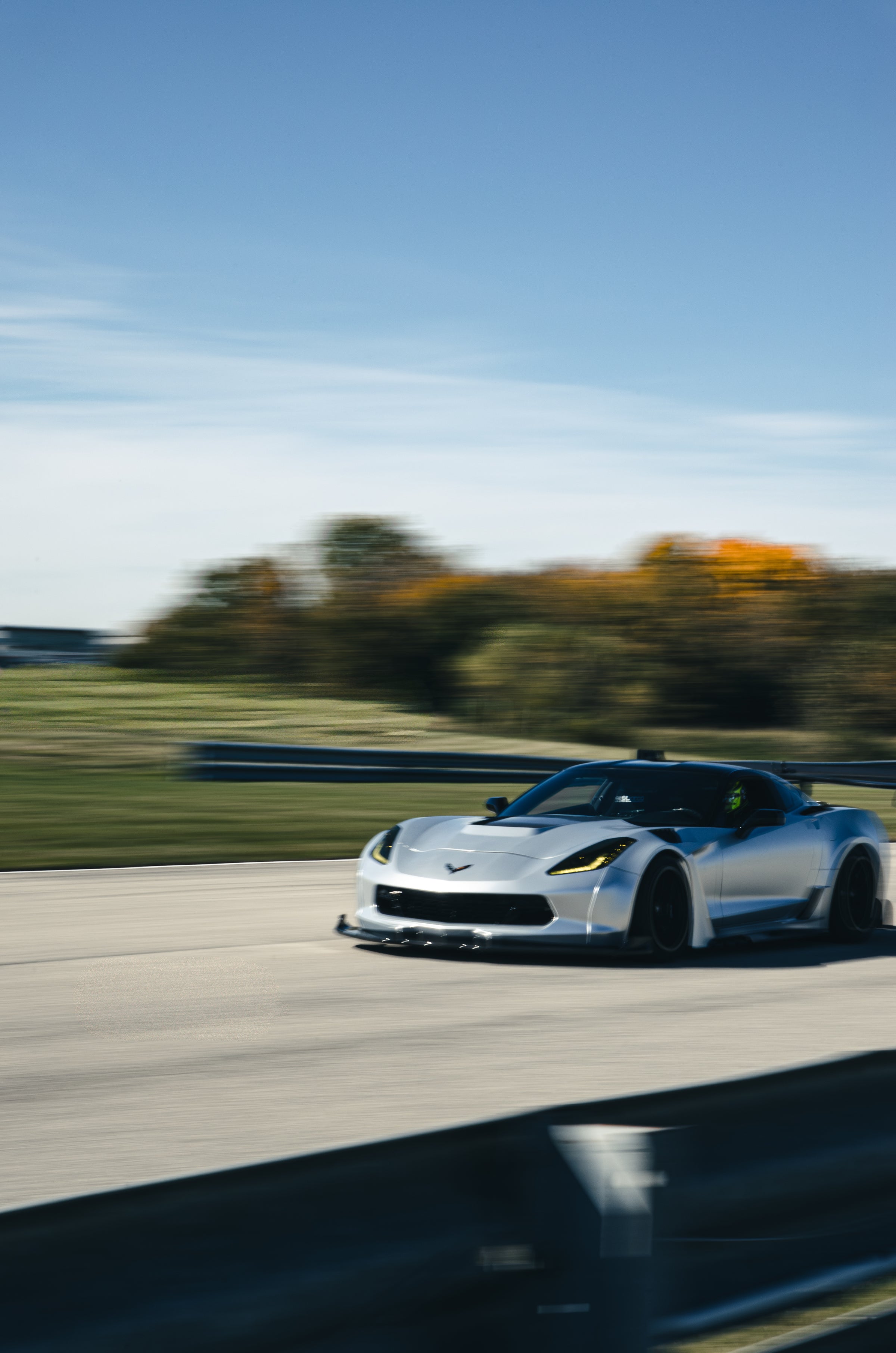 Silver Chevrolet Corvette C7 with carbon fiber front splitter driving on a sunny road, surrounded by autumn trees and a clear blue sky.