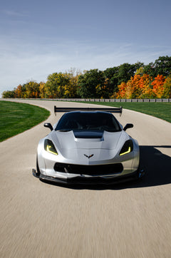 Silver Chevrolet Corvette C7 with carbon fiber front splitter driving on a sunny road, surrounded by autumn trees and a clear blue sky.