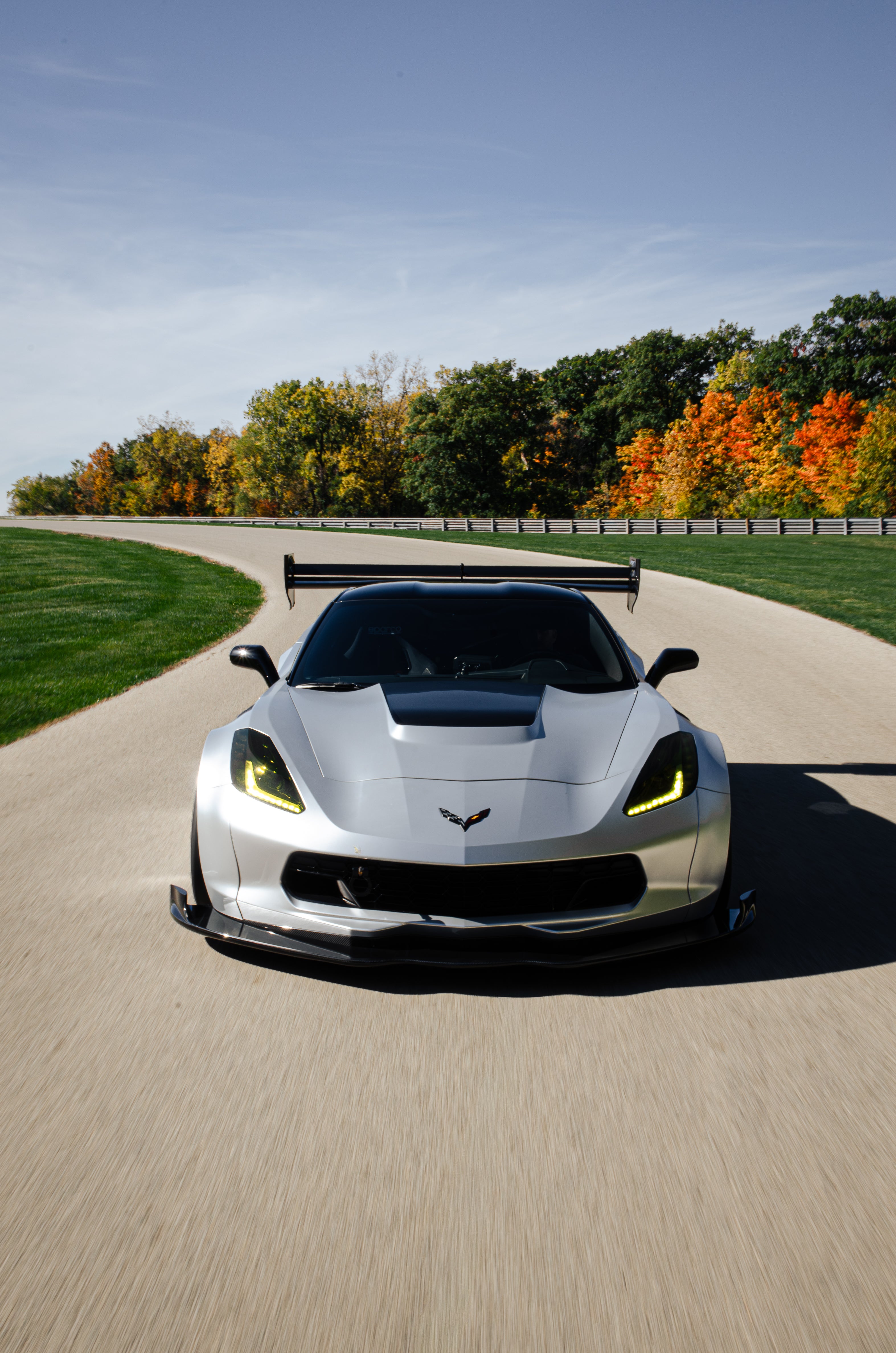 Silver Chevrolet Corvette C7 with carbon fiber front splitter driving on a sunny road, surrounded by autumn trees and a clear blue sky.