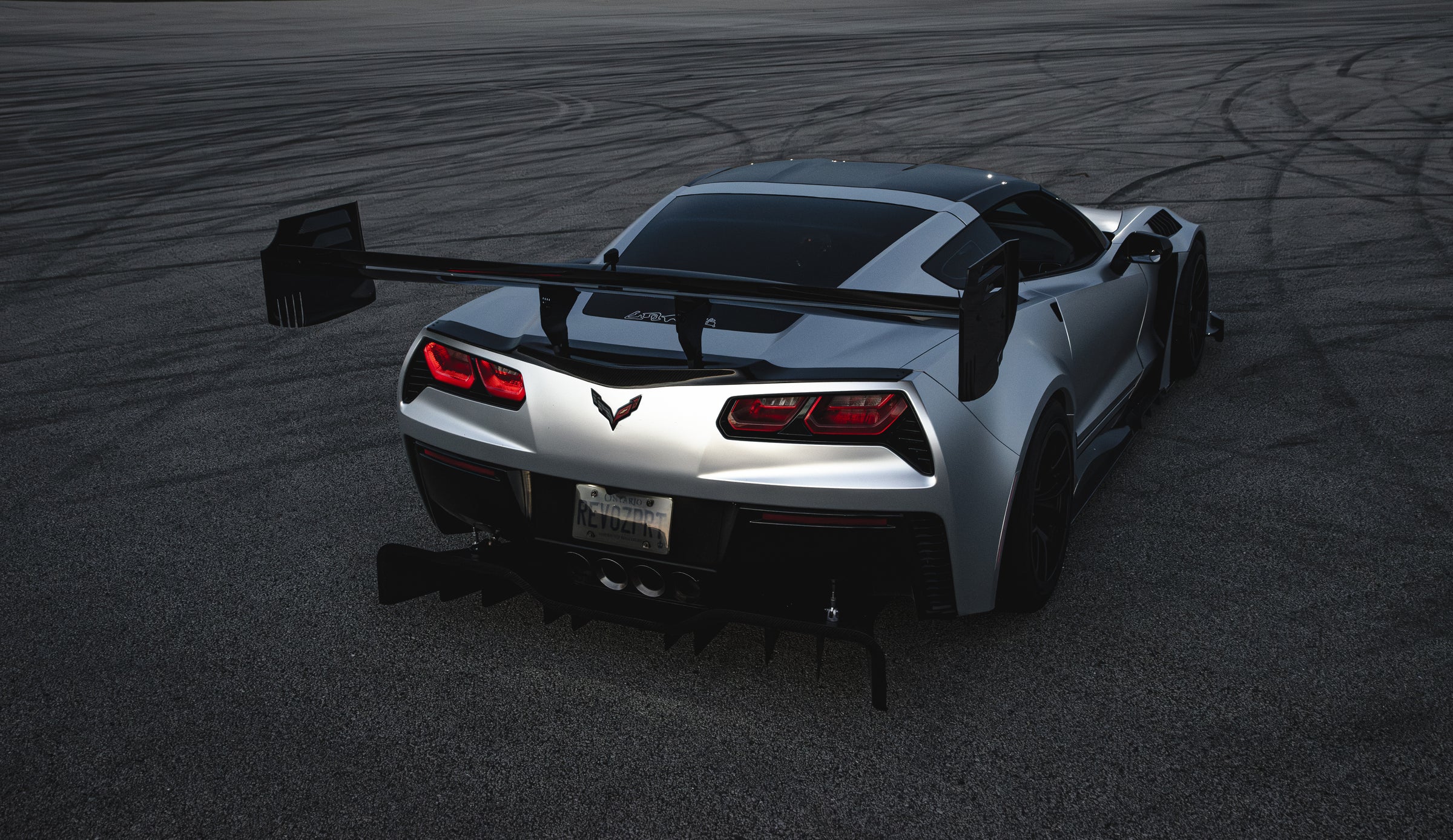 Silver Chevrolet Corvette C7 with carbon fiber front splitter driving on a sunny road, surrounded by autumn trees and a clear blue sky.