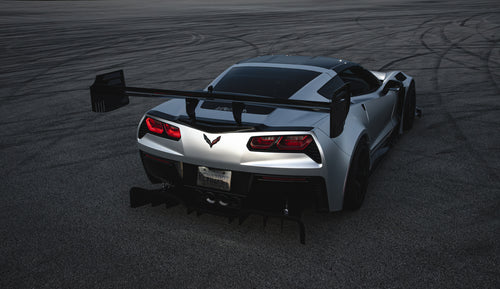 Silver Chevrolet Corvette C7 with carbon fiber front splitter driving on a sunny road, surrounded by autumn trees and a clear blue sky.