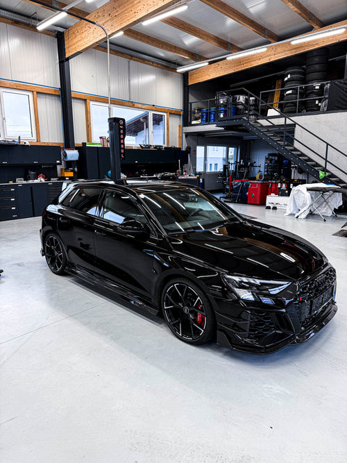 Black Audi RS3 Sportback featuring a REVOZPORT carbon fiber body kit with front lip and side skirts parked on a wet outdoor lot.