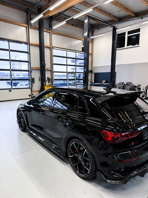 Black Audi RS3 Sportback featuring a REVOZPORT carbon fiber body kit with front lip and side skirts parked on a wet outdoor lot.