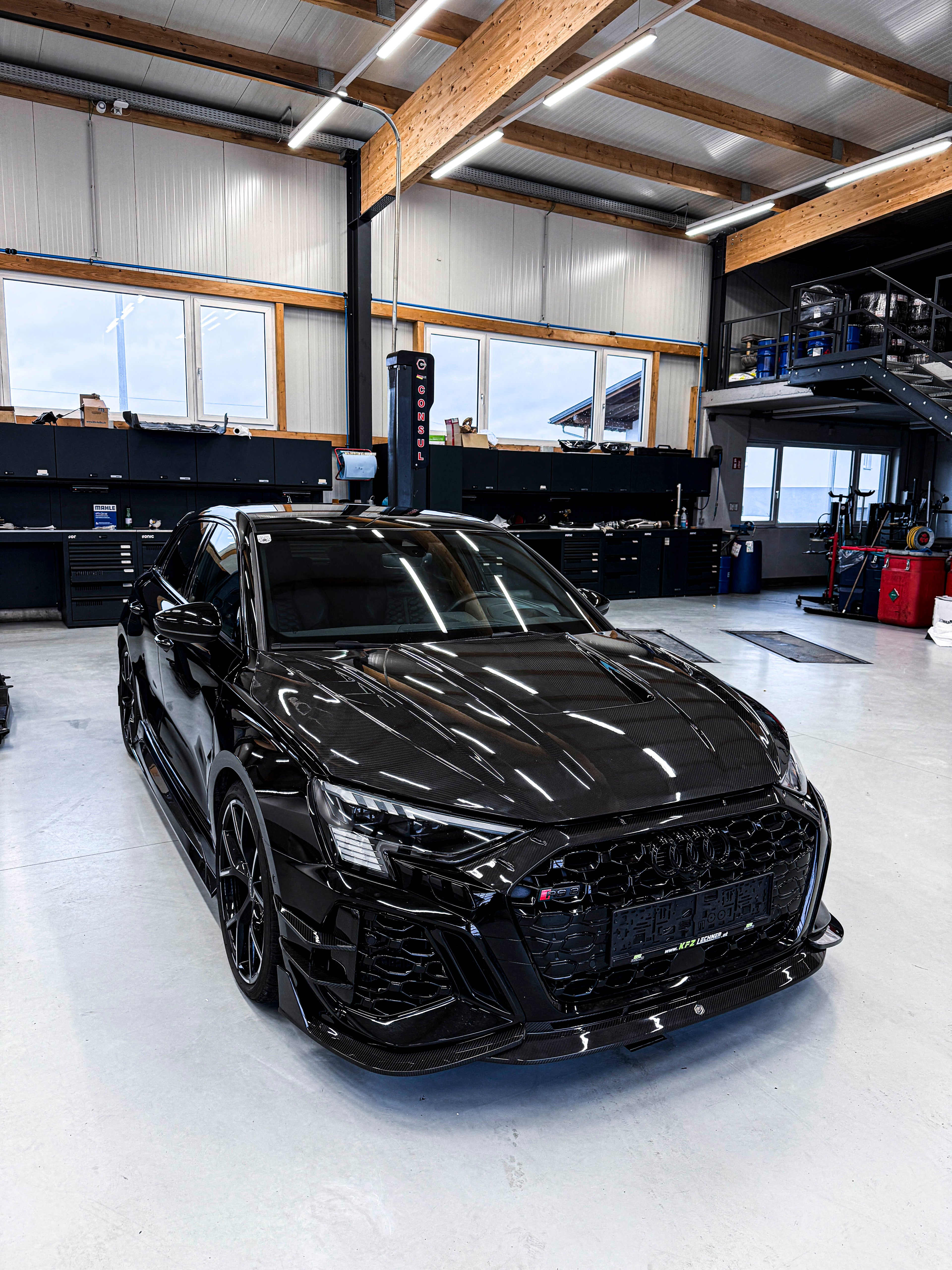 Black Audi RS3 Sportback featuring a REVOZPORT carbon fiber body kit with front lip and side skirts parked on a wet outdoor lot.