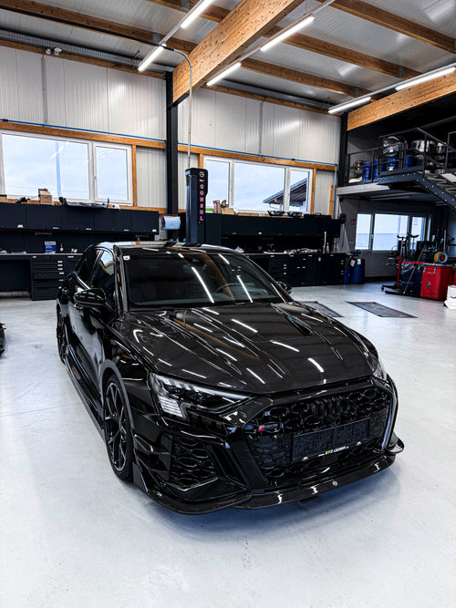 Black Audi RS3 Sportback featuring a REVOZPORT carbon fiber body kit with front lip and side skirts parked on a wet outdoor lot.