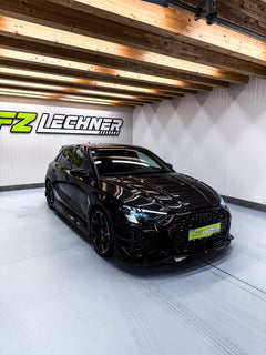Black Audi RS3 Sportback featuring a REVOZPORT carbon fiber body kit with front lip and side skirts parked on a wet outdoor lot.