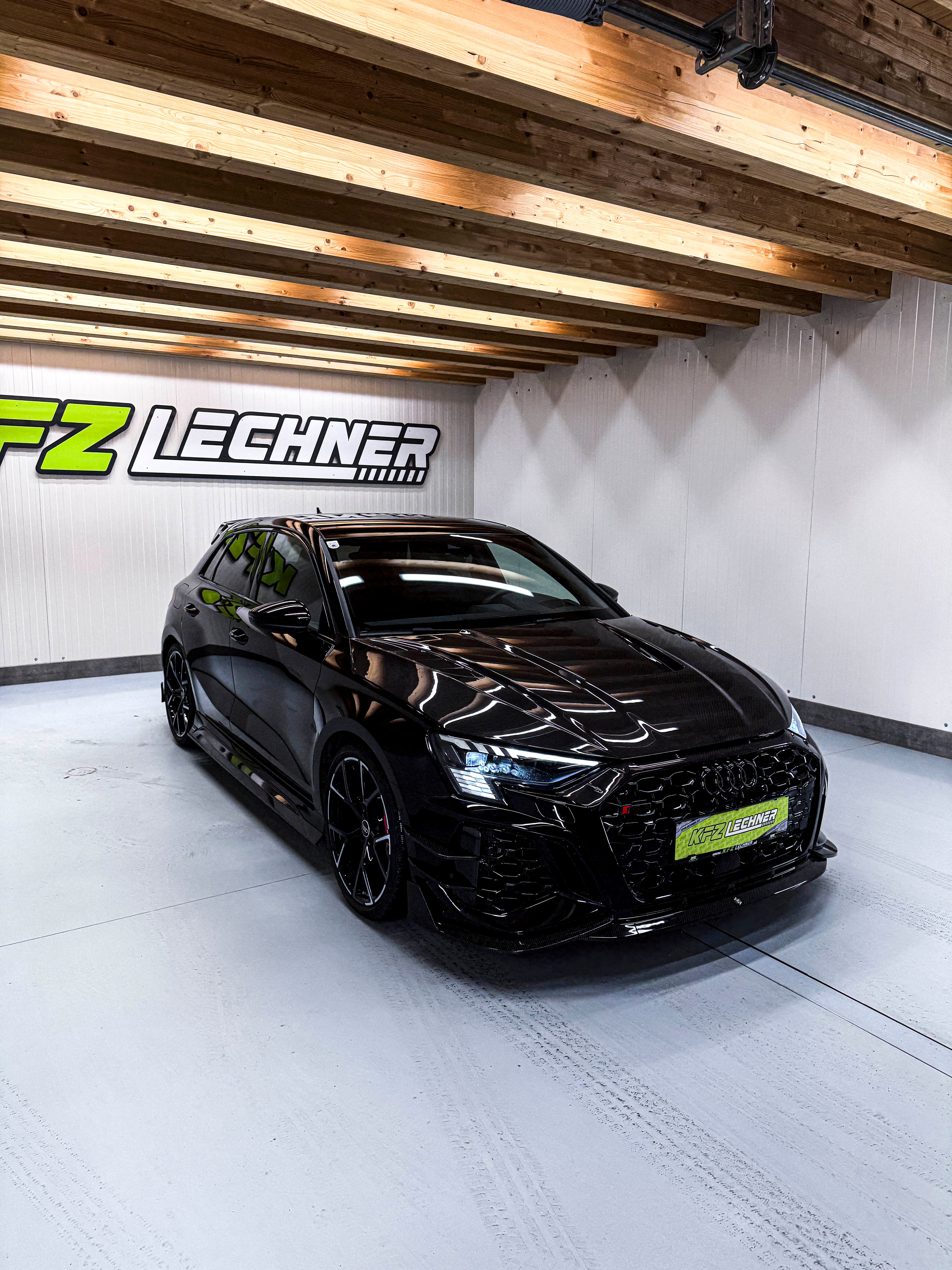 Black Audi RS3 Sportback featuring a REVOZPORT carbon fiber body kit with front lip and side skirts parked on a wet outdoor lot.