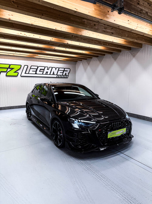 Black Audi RS3 Sportback featuring a REVOZPORT carbon fiber body kit with front lip and side skirts parked on a wet outdoor lot.