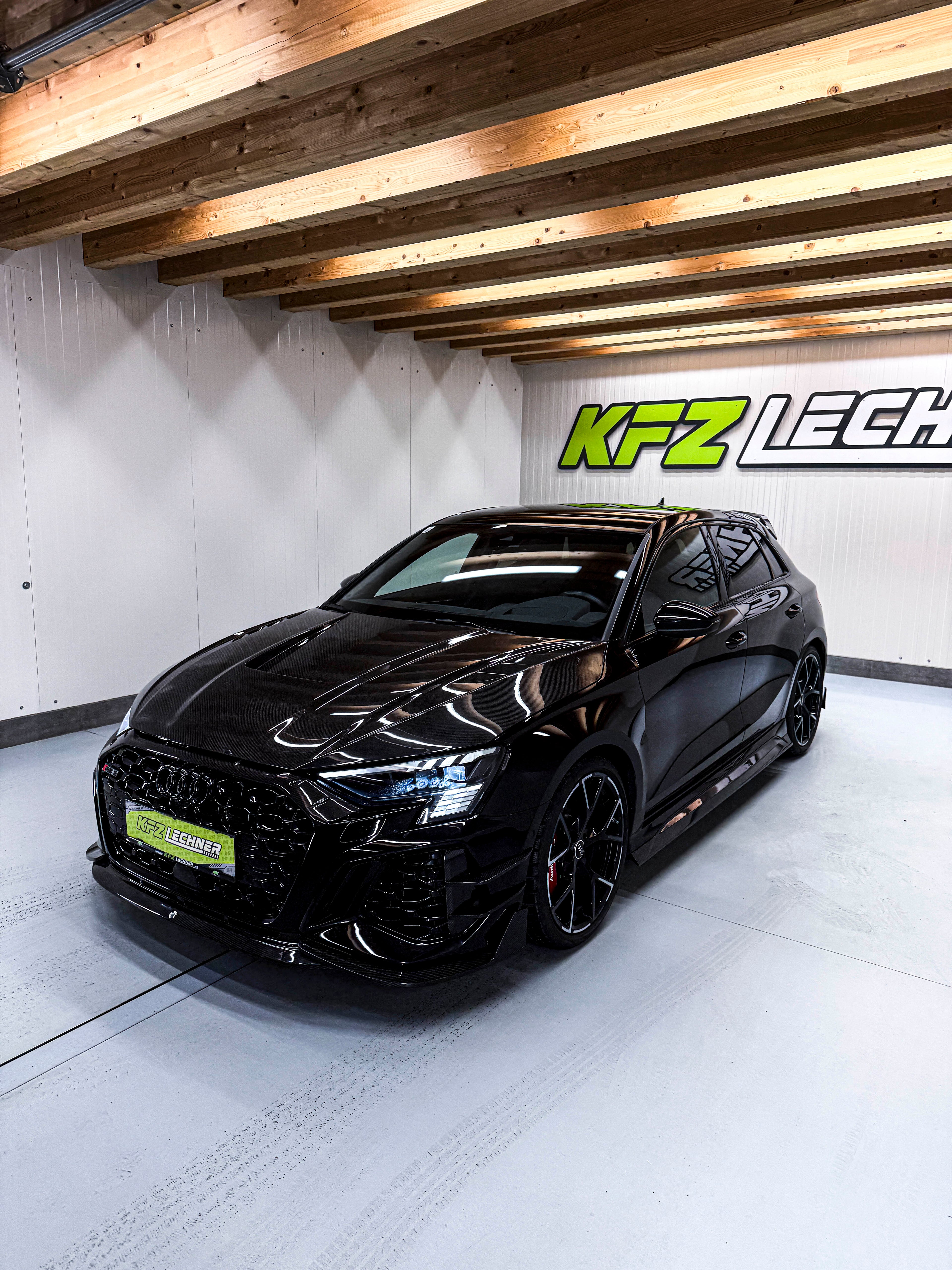 Black Audi RS3 Sportback featuring a REVOZPORT carbon fiber body kit with front lip and side skirts parked on a wet outdoor lot.