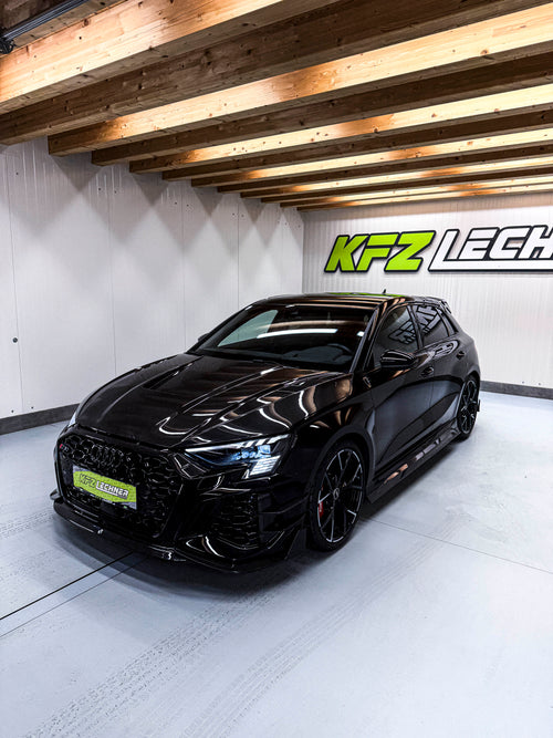 Black Audi RS3 Sportback featuring a REVOZPORT carbon fiber body kit with front lip and side skirts parked on a wet outdoor lot.
