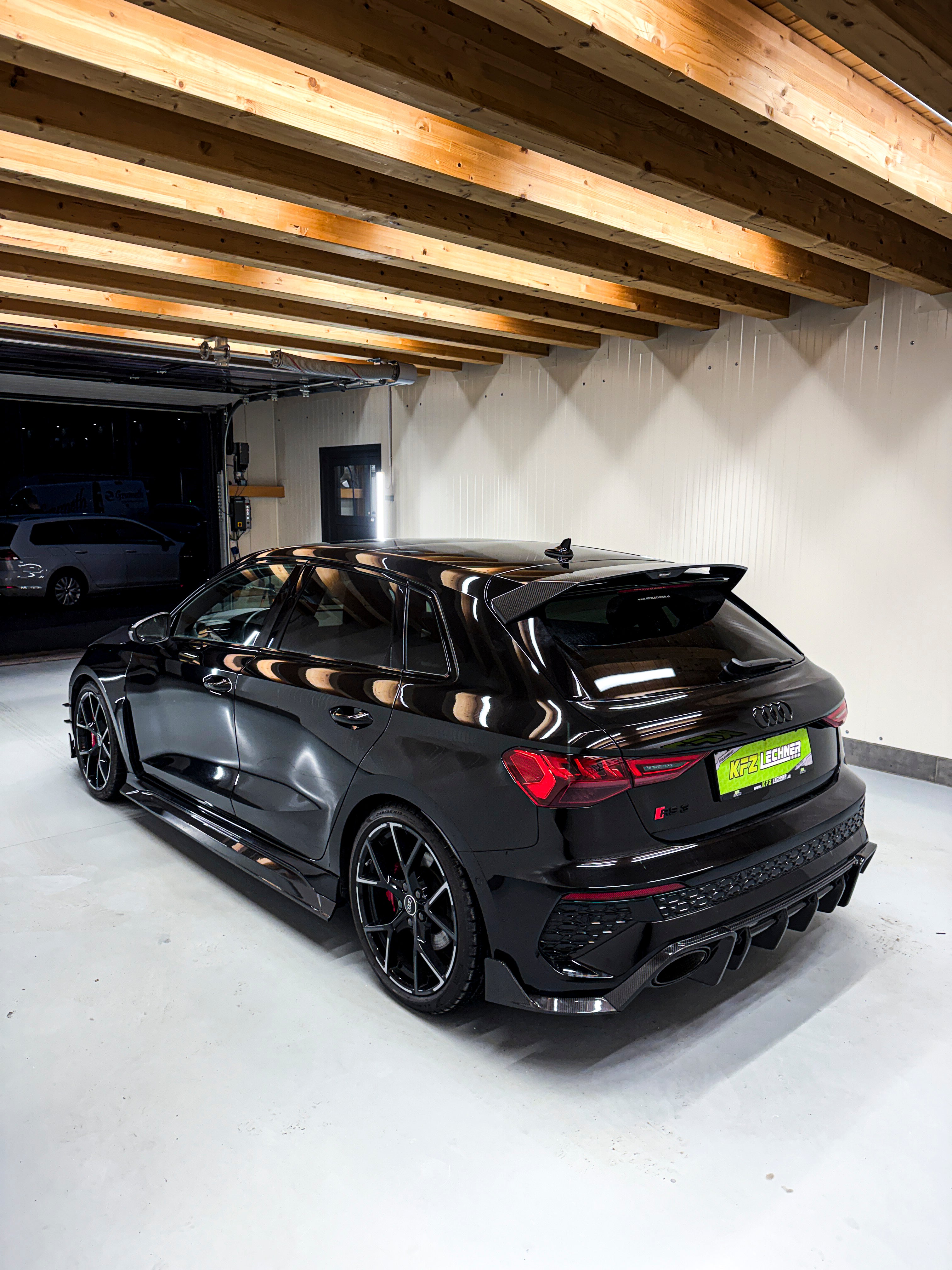 Black Audi RS3 Sportback featuring a REVOZPORT carbon fiber body kit with front lip and side skirts parked on a wet outdoor lot.