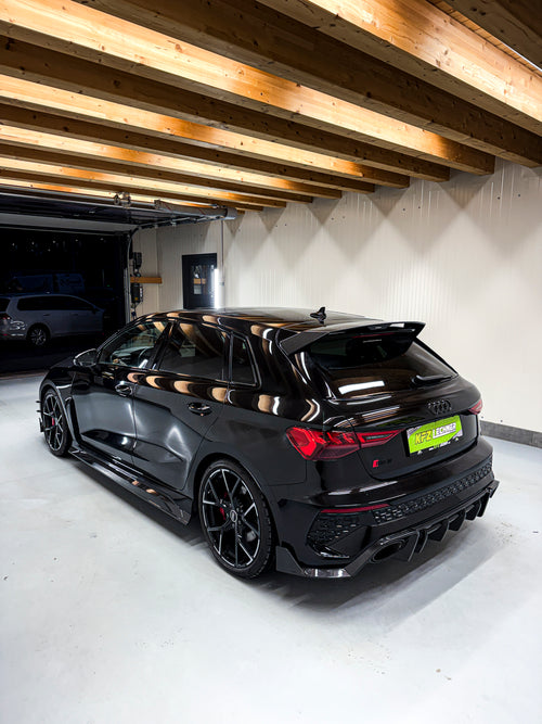 Black Audi RS3 Sportback featuring a REVOZPORT carbon fiber body kit with front lip and side skirts parked on a wet outdoor lot.
