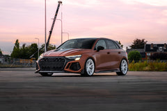 Bronze Audi RS3 Sportback featuring a REVOZPORT carbon fiber body kit with front lip and side skirts, paired with white multi-spoke forged wheels, parked at sunset in an open urban setting.