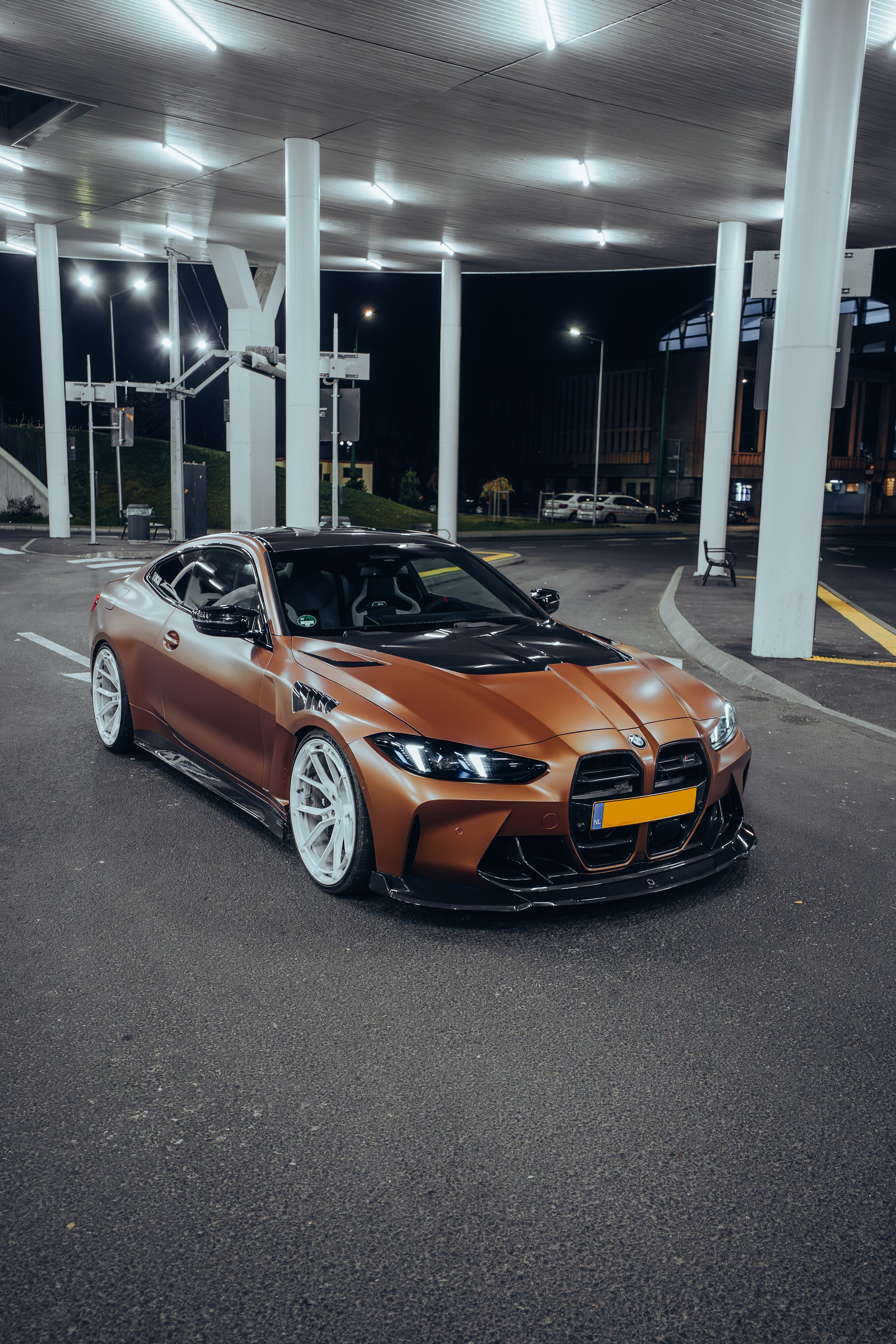 Bronze BMW G82 M4 featuring a REVOZPORT carbon fiber body kit with front lip, vented hood, side skirts, and white forged wheels parked under bright canopy lights at night.