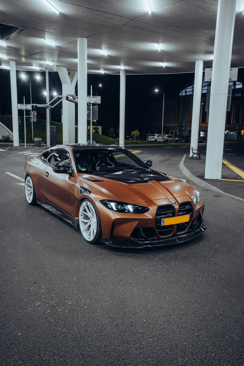 Bronze BMW G82 M4 featuring a REVOZPORT carbon fiber body kit with front lip, vented hood, side skirts, and white forged wheels parked under bright canopy lights at night.