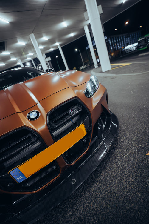 Bronze BMW G82 M4 featuring a REVOZPORT carbon fiber body kit with front lip, vented hood, side skirts, and white forged wheels parked under bright canopy lights at night.