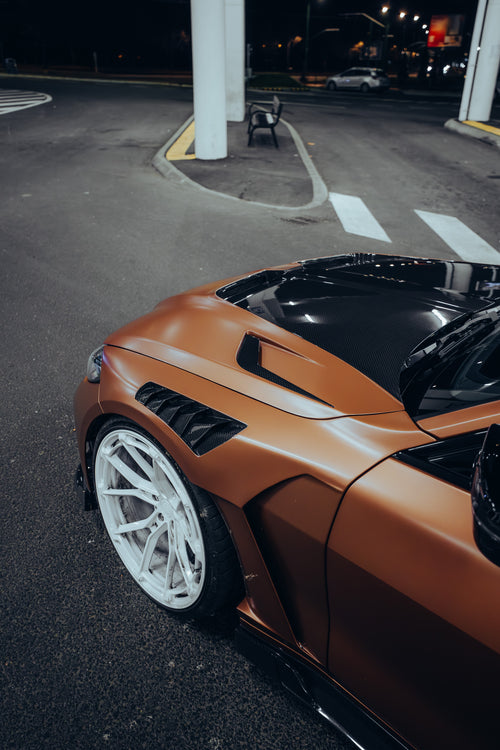 Bronze BMW G82 M4 featuring a REVOZPORT carbon fiber body kit with front lip, vented hood, side skirts, and white forged wheels parked under bright canopy lights at night.