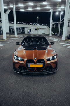 Bronze BMW G82 M4 featuring a REVOZPORT carbon fiber body kit with front lip, vented hood, side skirts, and white forged wheels parked under bright canopy lights at night.