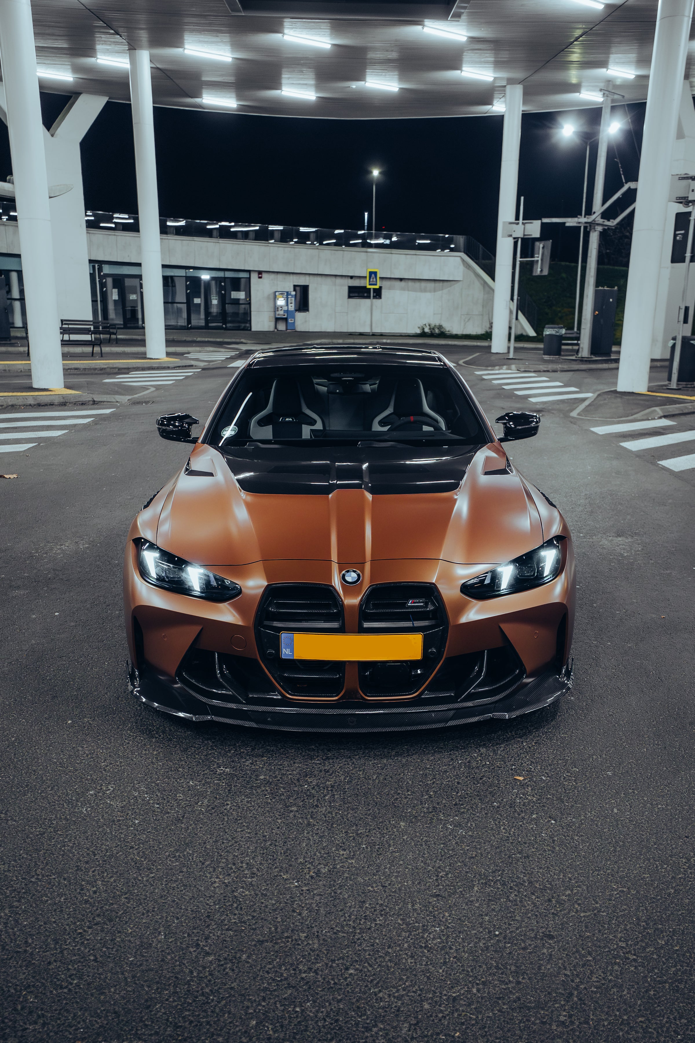 Bronze BMW G82 M4 featuring a REVOZPORT carbon fiber body kit with front lip, vented hood, side skirts, and white forged wheels parked under bright canopy lights at night.