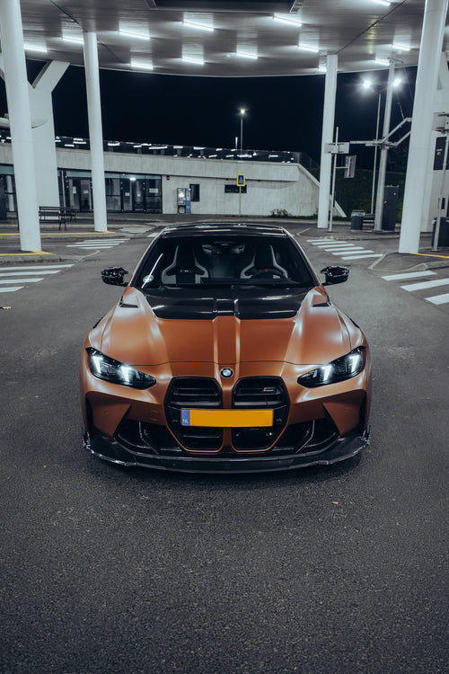 Bronze BMW G82 M4 featuring a REVOZPORT carbon fiber body kit with front lip, vented hood, side skirts, and white forged wheels parked under bright canopy lights at night.
