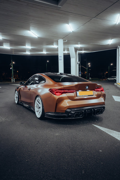 Bronze BMW G82 M4 featuring a REVOZPORT carbon fiber body kit with front lip, vented hood, side skirts, and white forged wheels parked under bright canopy lights at night.