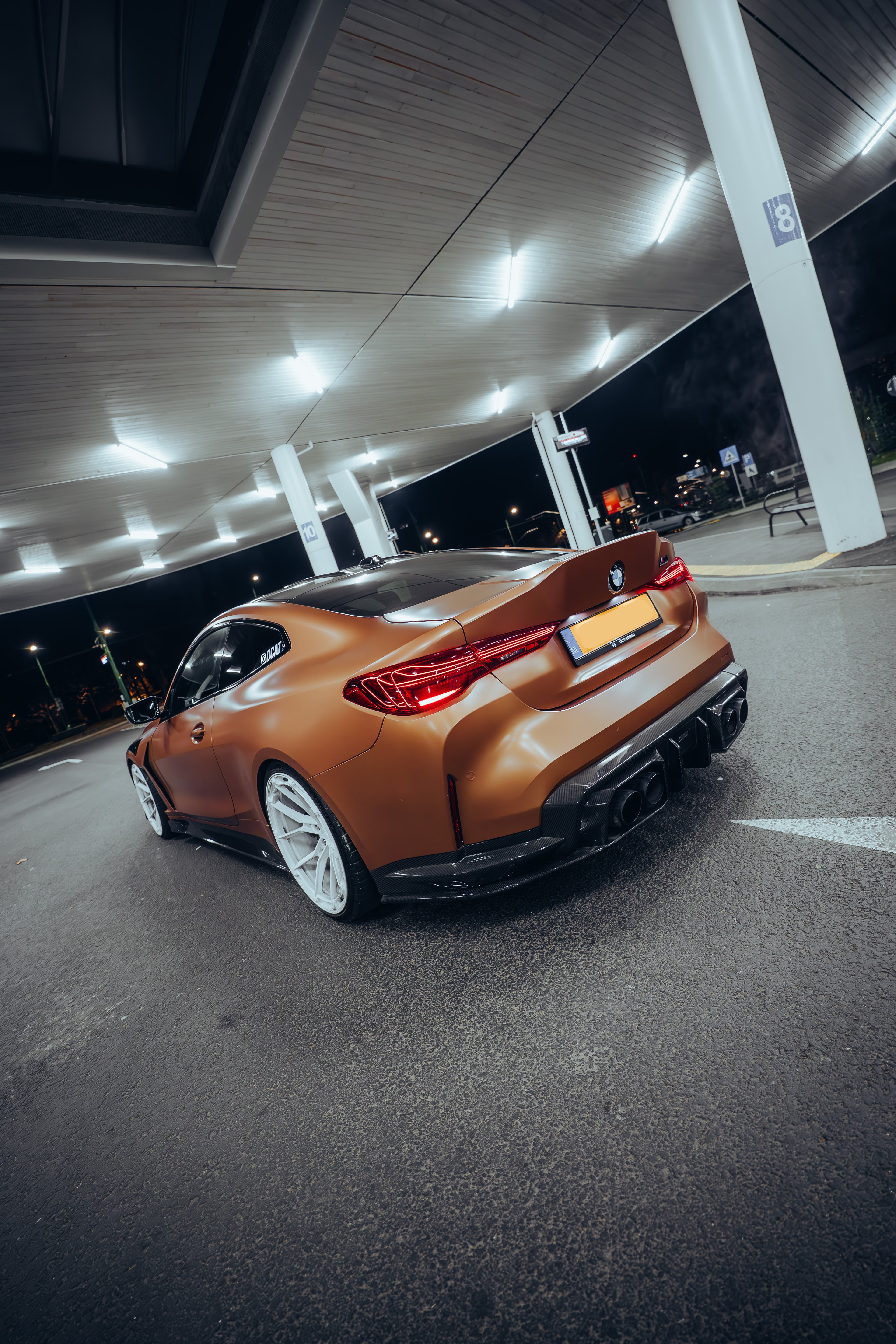 Bronze BMW G82 M4 featuring a REVOZPORT carbon fiber body kit with front lip, vented hood, side skirts, and white forged wheels parked under bright canopy lights at night.