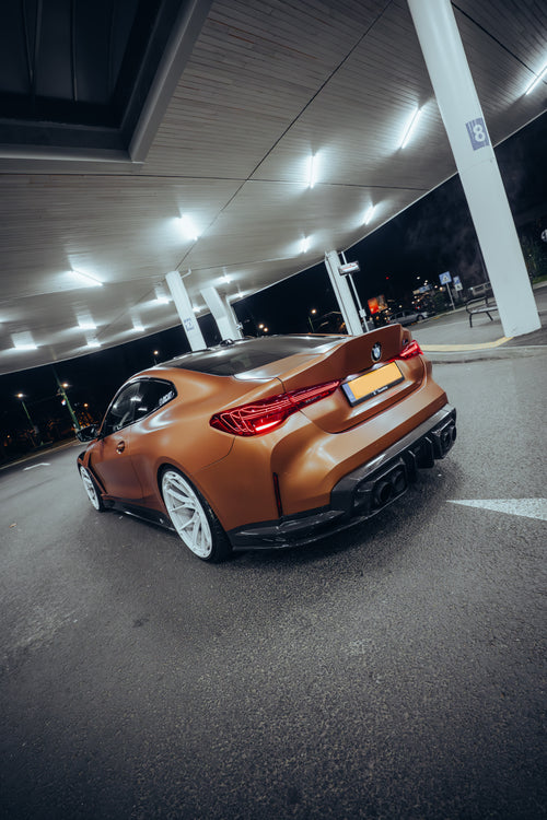 Bronze BMW G82 M4 featuring a REVOZPORT carbon fiber body kit with front lip, vented hood, side skirts, and white forged wheels parked under bright canopy lights at night.