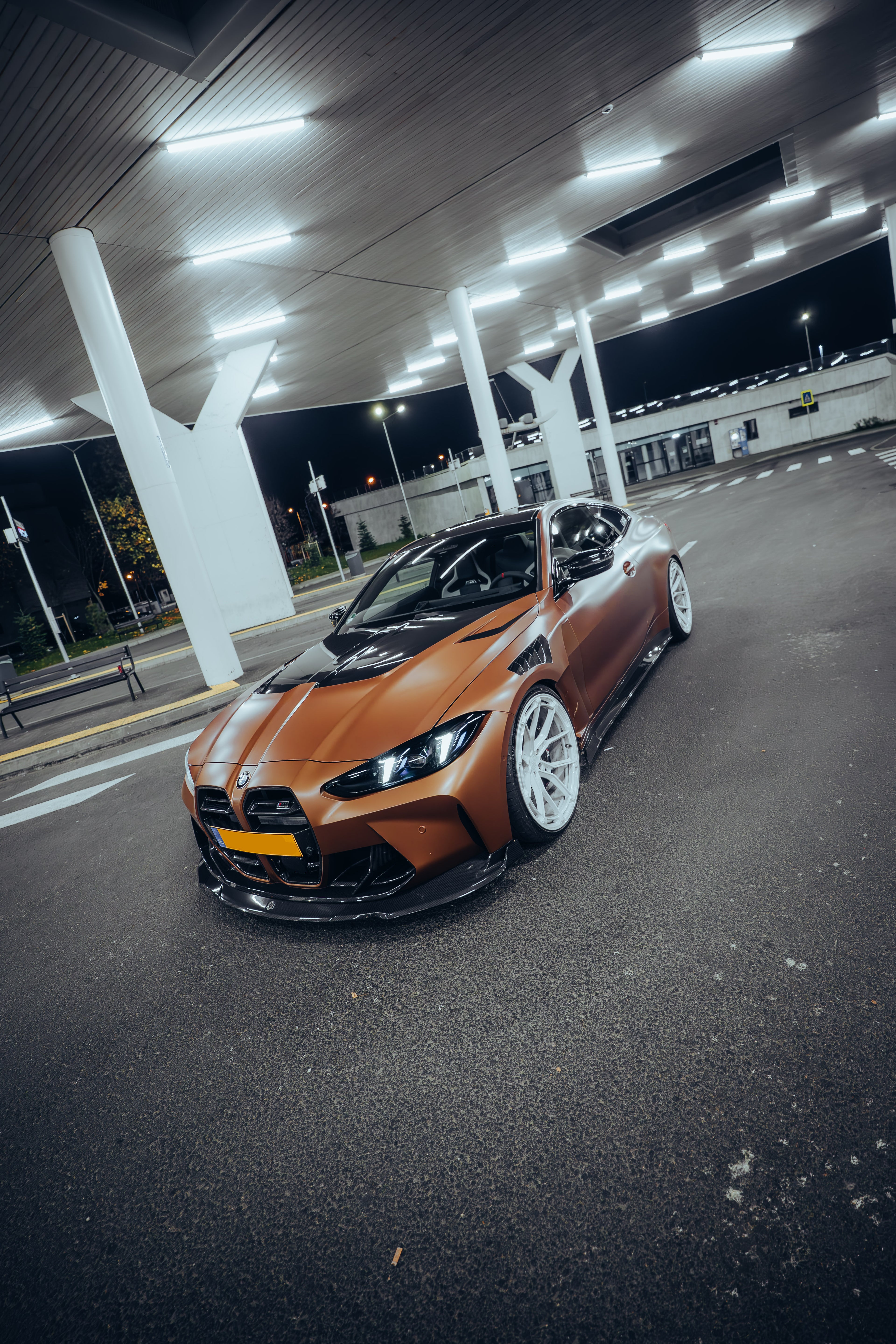 Bronze BMW G82 M4 featuring a REVOZPORT carbon fiber body kit with front lip, vented hood, side skirts, and white forged wheels parked under bright canopy lights at night.
