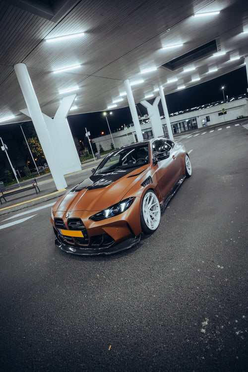 Bronze BMW G82 M4 featuring a REVOZPORT carbon fiber body kit with front lip, vented hood, side skirts, and white forged wheels parked under bright canopy lights at night.