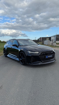 Black Audi RS6 Avant featuring a REVOZPORT carbon fiber body kit with front lip, side skirts, aggressive aero styling, and blacked-out wheels parked on an open road.