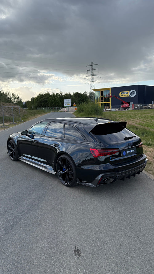 Black Audi RS6 Avant featuring a REVOZPORT carbon fiber body kit with front lip, side skirts, aggressive aero styling, and blacked-out wheels parked on an open road.