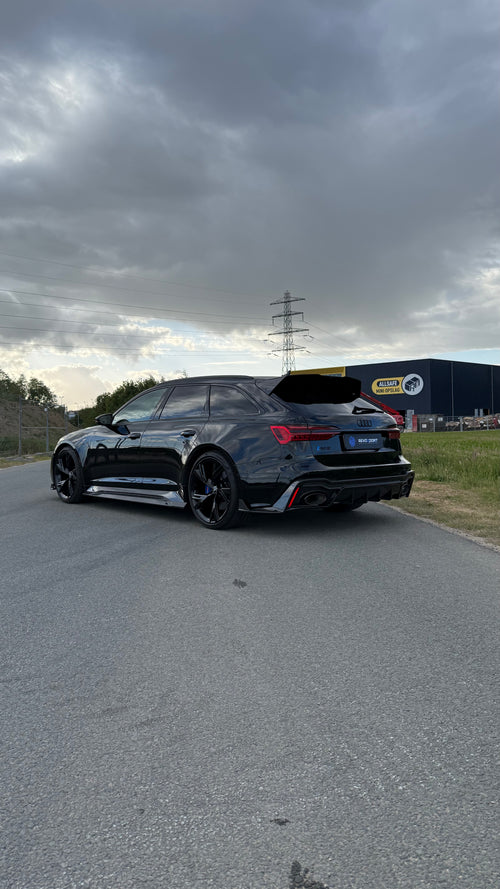 Black Audi RS6 Avant featuring a REVOZPORT carbon fiber body kit with front lip, side skirts, aggressive aero styling, and blacked-out wheels parked on an open road.