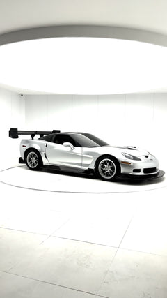 Close-up of a silver wide-body Chevrolet Corvette performance car displayed indoors, featuring aggressive aerodynamic bodywork, carbon fiber front splitter, vented hood, and lightweight multi-spoke wheels. The image highlights the custom motorsport styling and premium carbon fiber aero components under exhibition lighting.
