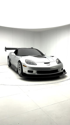 Close-up of a silver wide-body Chevrolet Corvette performance car displayed indoors, featuring aggressive aerodynamic bodywork, carbon fiber front splitter, vented hood, and lightweight multi-spoke wheels. The image highlights the custom motorsport styling and premium carbon fiber aero components under exhibition lighting.