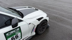 White BMW G80 M3 race car with REVOZPORT carbon fiber aero kit, front splitter, canards, and large rear wing on a wet track paddock during a motorsport event.