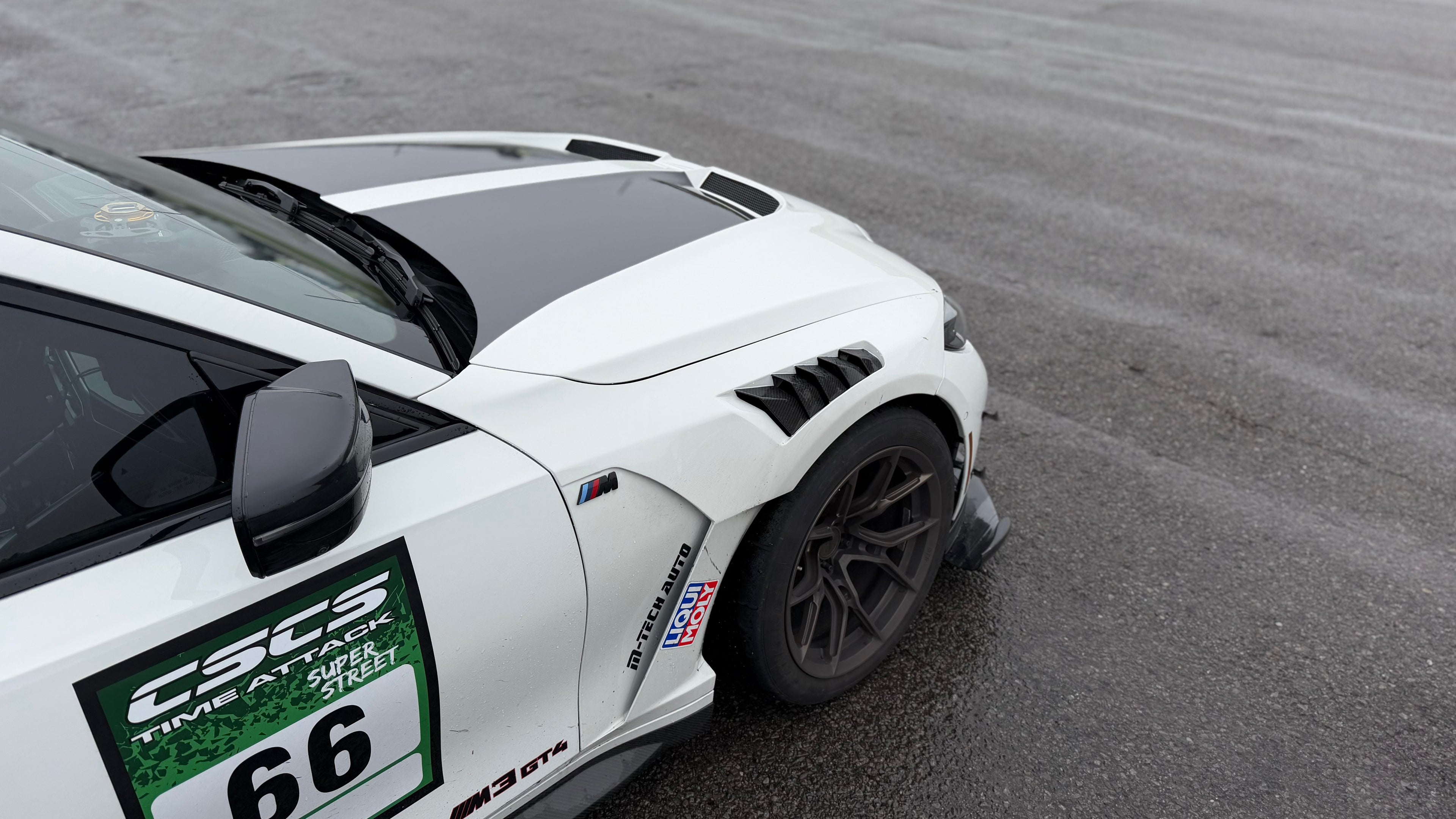 White BMW G80 M3 race car with REVOZPORT carbon fiber aero kit, front splitter, canards, and large rear wing on a wet track paddock during a motorsport event.