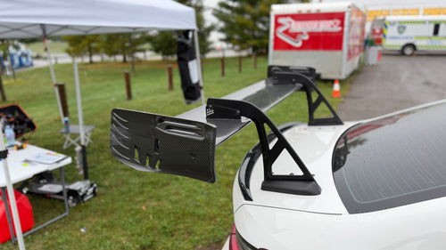 White BMW G80 M3 race car with REVOZPORT carbon fiber aero kit, front splitter, canards, and large rear wing on a wet track paddock during a motorsport event.