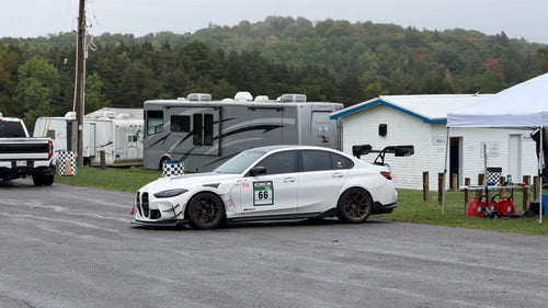 White BMW G80 M3 race car with REVOZPORT carbon fiber aero kit, front splitter, canards, and large rear wing on a wet track paddock during a motorsport event.