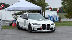 White BMW G80 M3 race car with REVOZPORT carbon fiber aero kit, front splitter, canards, and large rear wing on a wet track paddock during a motorsport event.